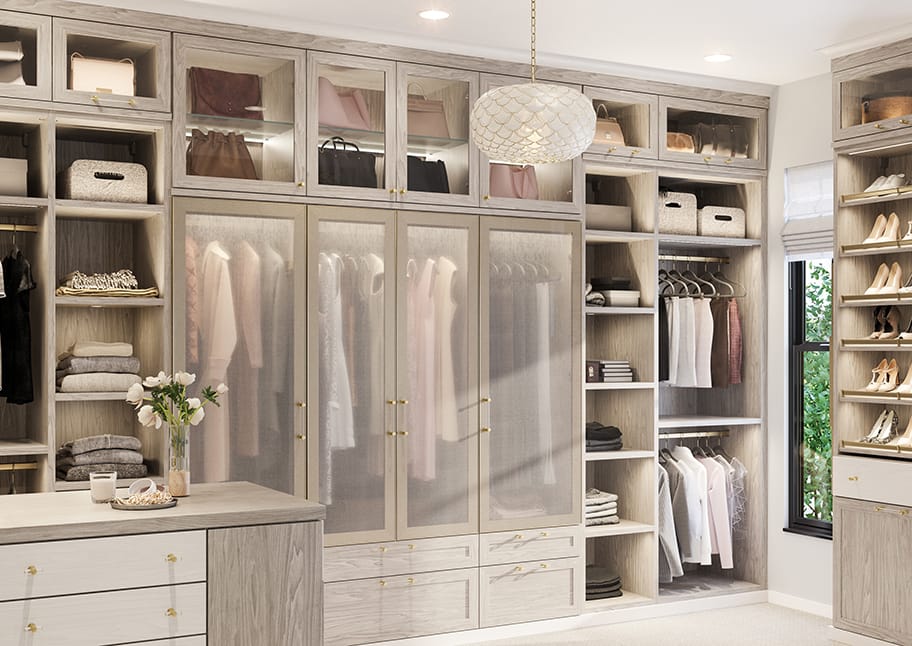 Walk in closet designed with glass cabinet doors, vanity, island and shoe storage by California Closets